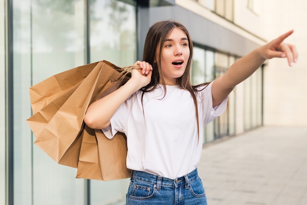 Acquirente stupito che apre la bocca tenendo le borse della spesa guardando le offerte speciali nei negozi e indicando la strada