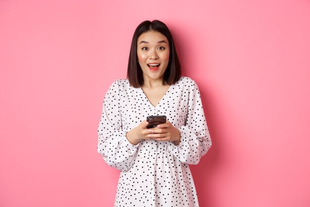 Acquisti online. Donna asiatica stupita che guarda la telecamera con un sorriso felice, fa acquisti con lo smartphone, utilizza l'app del telefono cellulare, in piedi su sfondo rosa
