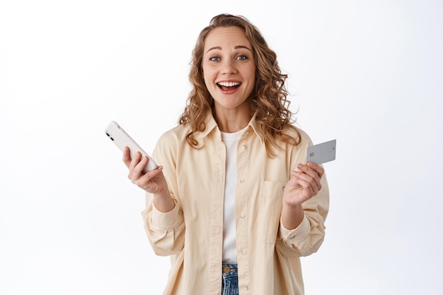 Acquisti online Giovane donna bionda che paga con carta di credito in smartphone sorridente e guardando felice sfondo bianco