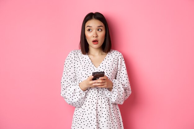 Acquisti online. Ragazza asiatica confusa che tiene in mano uno smartphone, guardando all'oscuro nell'angolo in alto a sinistra, in piedi su sfondo rosa.