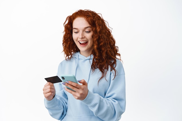 Acquisti online. Ritratto di donna dai capelli rossi che paga in internet, con in mano una carta di credito in plastica e uno smartphone, in piedi su bianco