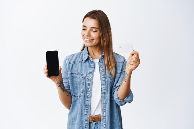 Acquisti online. Ritratto di giovane donna che fa soldi con l'app, mostrando lo schermo vuoto dello smartphone e la carta di credito in plastica sorridente, guardando l'applicazione mobile, bianco