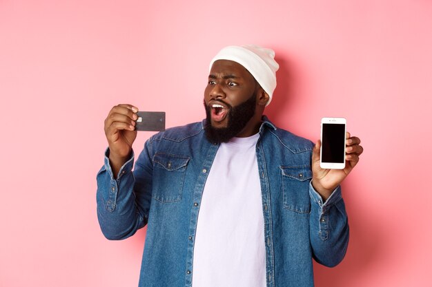 Acquisti online. Uomo di colore scioccato che mostra lo schermo del telefono cellulare, guardando sorpreso la carta di credito, in piedi in abiti hipster su sfondo rosa.