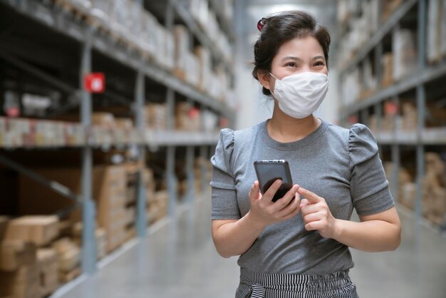 Acquisto casuale asiatico femminile indossare maschera viso uso mano smartphone assistenza allo shopping utilizzando l'applicazione per il controllo delle scorte e la descrizione della quantità per un facile acquisto nella sala dell'inventario del magazzino