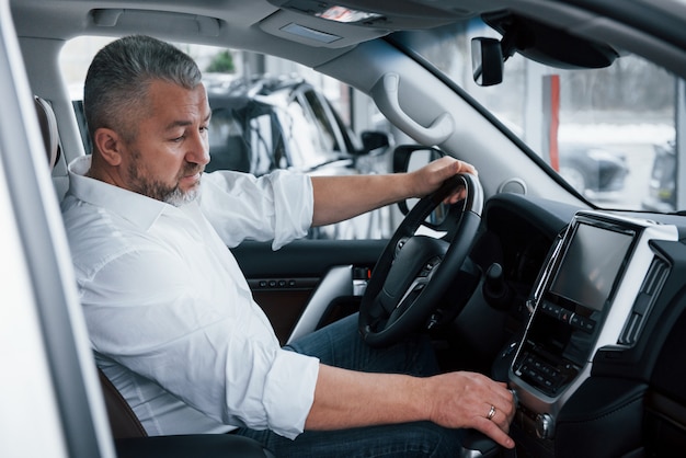 Acquisto e test di auto nuove. L'uomo d'affari senior in vestiti ufficiali si siede in un'auto di lusso e spingendo i bottoni sul lettore musicale