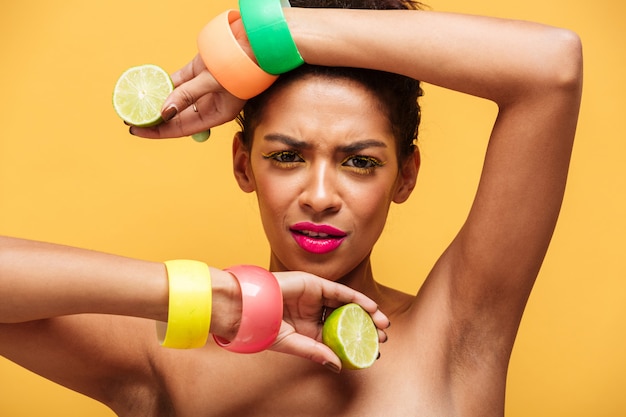 Adatti il ritratto della donna afroamericana con trucco d'avanguardia e gli accessori che tengono le due metà di calce matura fresca in entrambe le mani isolate, sopra la parete gialla