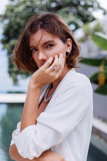 Adatti il ritratto della donna alla villa di lusso tropicale che porta blazer alla moda bianco e gioielli sopra le foglie tropicali