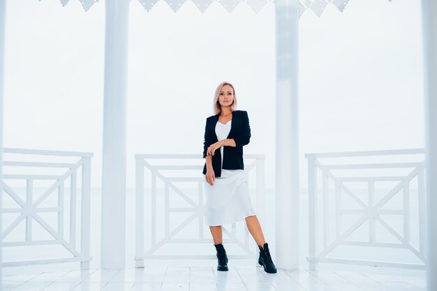 Adatti il ritratto della donna caucasica alla moda in blazer nero vestito lungo di seta e grandi stivali in luogo di lusso con vista sul mare