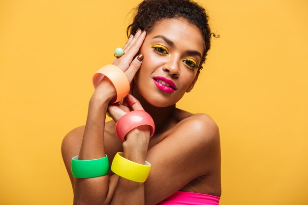 Adatti il ritratto di bella donna afroamericana con trucco luminoso che dimostra i gioielli multicolori che si tengono per mano al fronte isolato, sopra giallo