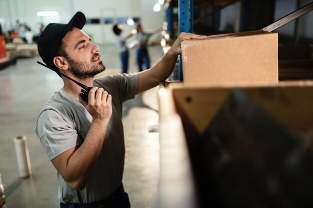 Addetto al magazzino che controlla i pacchi per la spedizione e utilizza il walkietalkie nel vano di stoccaggio industriale