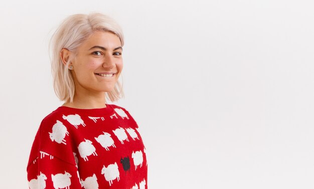 Adolescente alla moda dall'aspetto trendy con capelli tinti di rosa e anello al naso sorridente, vestito con un maglione rosso lavorato a maglia con stampa carina. Piuttosto giovane donna che si morde il labbro, con uno sguardo civettuolo