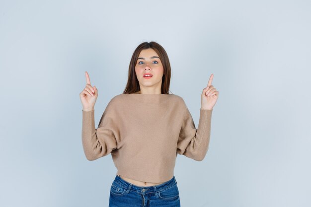 Adolescente che indica in su in maglione, jeans e che sembra astuto. vista frontale.