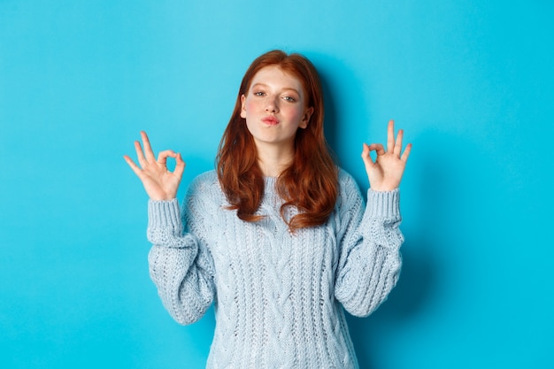 Adolescente dai capelli rossi che mostra segni ok, sembra soddisfatta e orgogliosa, d'accordo, dà una risposta positiva, lodando la buona scelta, in piedi su sfondo blu