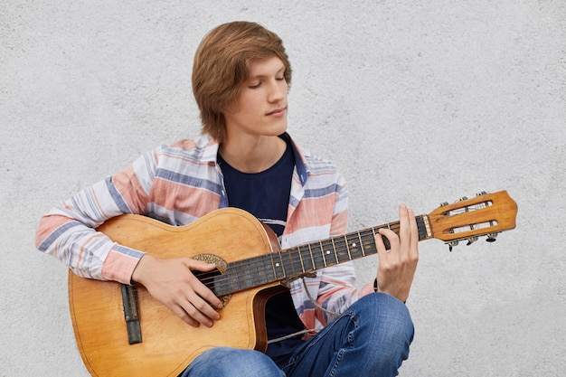 Adolescente di talento con la pettinatura alla moda che tiene la chitarra acustica suonando le sue canzoni preferite mentre era seduto contro il muro di cemento grigio