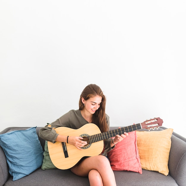 Adolescente felice che si siede sul sofà che gioca chitarra contro la priorità bassa bianca