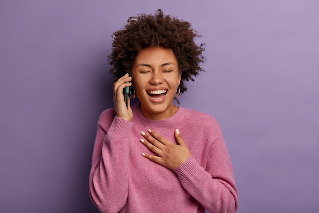 Adolescente felicissima ride durante le conversazioni telefoniche, tiene la mano sul petto, non riesce a smettere di ridacchiare, tiene il cellulare vicino all'orecchio, sente battute divertenti, chiude gli occhi, modella al chiuso contro il muro viola