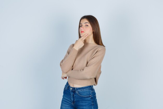 Adolescente in maglione, jeans che tiene le dita sulla mascella e sembra premurosa, vista frontale.