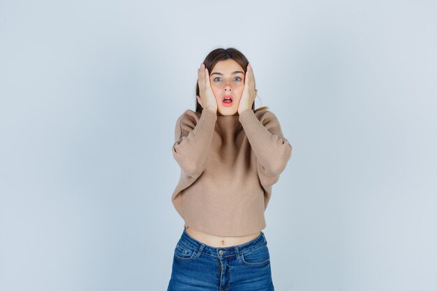 Adolescente in maglione, jeans che tiene le mani sulle guance e sembra ansiosa, vista frontale.