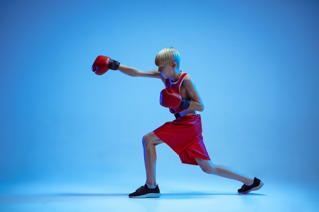 Adolescente nella boxe di abbigliamento sportivo isolato su sfondo blu studio in luce al neon. Pugile caucasico maschio novizio che si allena duro e lavora fuori, calci. Sport, stile di vita sano, concetto di movimento.