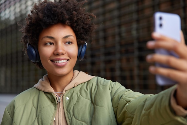 Adolescente positivo con i capelli ricci prende selfie tramite smartphone ascolta la traccia audio tramite le cuffie gode di passeggiate nel tempo libero all'aperto vestita con felpa con cappuccio e giacca pose su sfondo sfocato