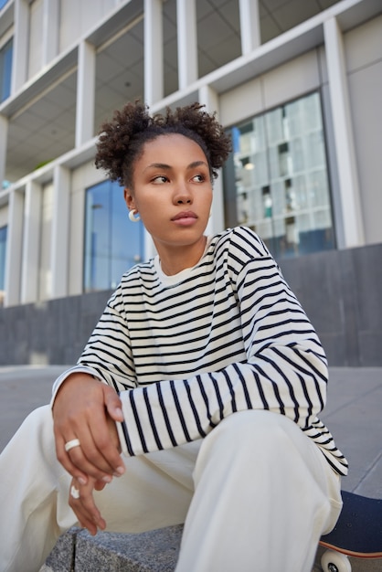 Adolescente premurosa vestita con maglione a righe e pantaloni distoglie lo sguardo posa sul longboard contempla qualcosa che posa contro l'edificio urbano ama lo sport estremo