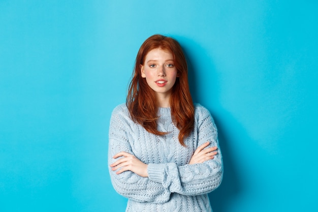 Adolescente rossa in maglione caldo in piedi su sfondo blu, braccia incrociate sul petto con fiducia.