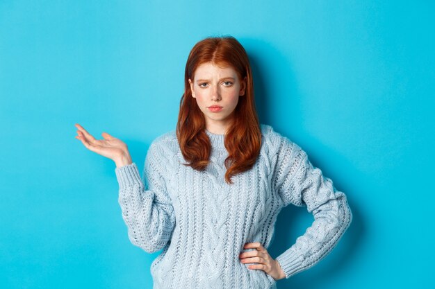 Adolescente scettica che sembra non divertita, alzando la mano in quel gesto, fissando qualcosa con una faccia distratta, in piedi su sfondo blu.