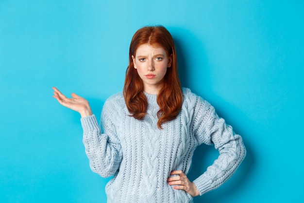 Adolescente scettica che sembra non divertita, alzando la mano in quel gesto, fissando qualcosa con una faccia distratta, in piedi su sfondo blu.