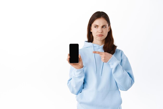 Adolescente triste delusa che punta il dito sullo schermo vuoto dello smartphone che mostra il display vuoto mobile e imbronciato sconvolto accigliato dispiaciuto in piedi su sfondo bianco