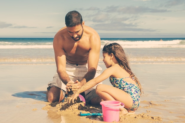 Adorabile bambina e suo padre che costruiscono castelli di sabbia sulla spiaggia, seduti sulla sabbia bagnata, godendo le vacanze