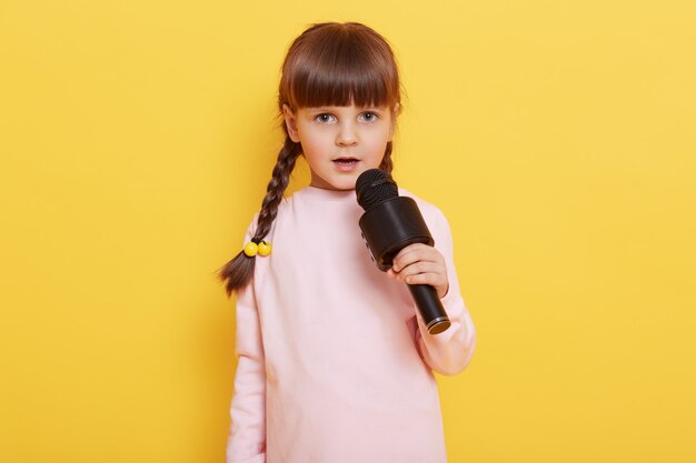 Adorabile bambino carino con il microfono nelle mani che canta canzoni, guarda la fotocamera, esibendosi isolato su sfondo giallo, bambino che organizza un concerto, canta in karaoke.