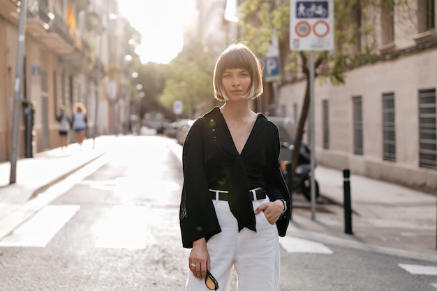 Adorabile donna europea adorabile con acconciatura corta che indossa abiti casual in posa davanti alla telecamera al sole sulla strada estiva in una calda giornata di sole