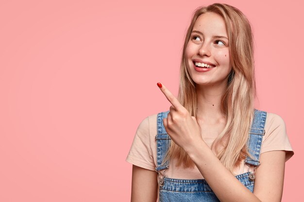 Adorabile giovane donna allegra con un sorriso positivo, punti da parte, mostra uno spazio vuoto, essendo di alto spirito, vestita con abiti casual