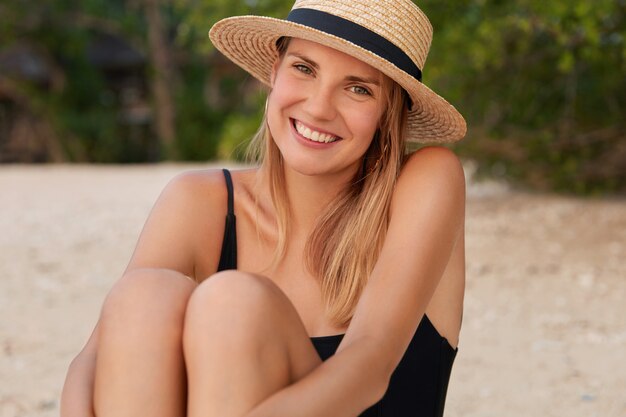 Adorabile giovane donna con un'espressione soddisfatta, prende il sole sulla spiaggia, si siede sulla spiaggia sabbiosa, indossa un cappello di paglia estivo e un costume da bagno nero, ha la pelle abbronzata.