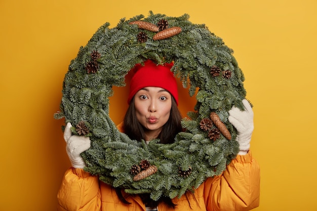 Adorabile giovane donna coreana mantiene le labbra arrotondate, guarda attraverso la corona di abete verde, indossa un cappotto giallo e guanti bianchi, decora la casa prima di Natale, posa al coperto.