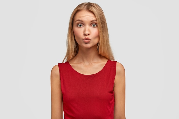 Adorabile giovane donna tiene le labbra arrotondate, va a baciare il suo ragazzo, apre gli occhi ampiamente, vestita con abiti rossi alla moda