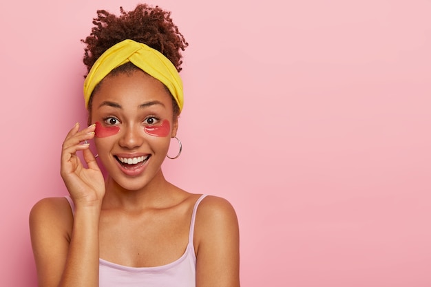 adorabile modella dalla pelle scura con capelli croccanti, applica macchie di idrogel rosa sotto gli occhi per rimuovere borse e gonfiori dopo una notte insonne, ha un'espressione allegra, vestita in modo casual