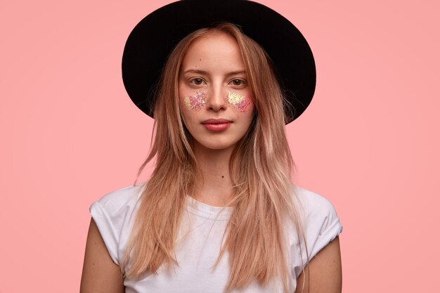 Adorabile modella giovane donna europea con glitter sul viso, indossa un elegante cappello nero, maglietta bianca, posa sul muro rosa, pronta per il festival con gli amici