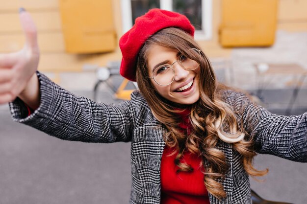 Adorabile modello femminile dai capelli scuri in occhiali alla moda che fa selfie vicino all'edificio giallo. Ritratto del primo piano della ragazza carina raffinata in cappello rosso che si diverte all'aperto.