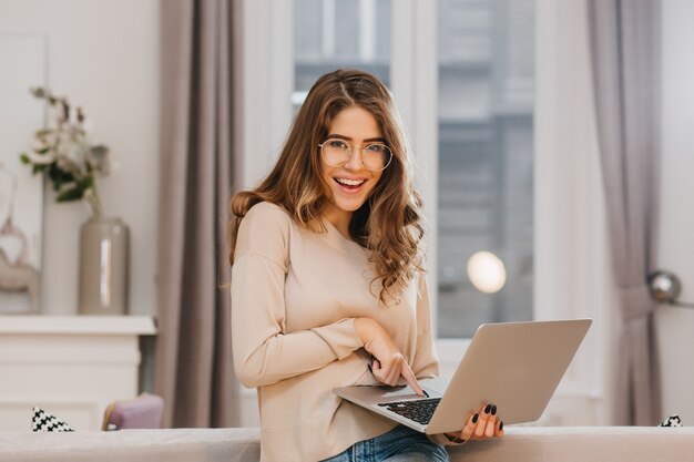 Adorabile professionista femminile in occhiali alla moda in posa con piacere durante il lavoro con il computer portatile