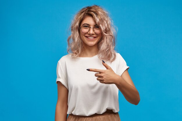 Adorabile ragazza alla moda caucasica hipster in abiti alla moda e accessori con un sorriso carino cheerul fiducioso, puntando il dito indice lateralmente