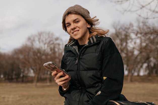 Adorabile ragazza amichevole che indossa un berretto lavorato a maglia e un giubbotto scuro sta riposando all'aperto contro il parco con lo smartphone