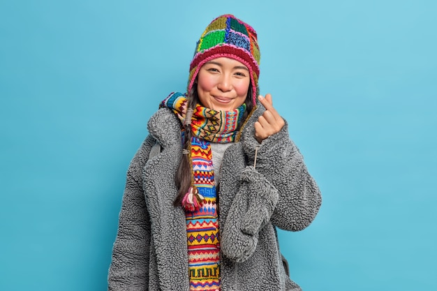 Adorabile ragazza asiatica orientale vestita in abiti invernali rende coreano come il segno mostra mini cuore esprime amore andando a fare pose a piedi contro il muro blu dello studio