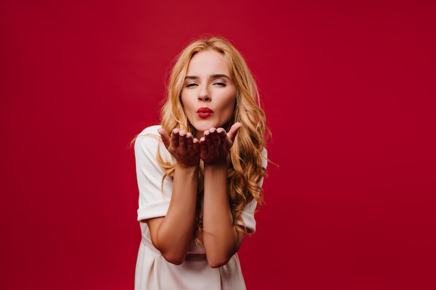 Adorabile ragazza bionda che manda baci d'aria durante il servizio fotografico. Carina donna europea in abito bianco che esprime amore.