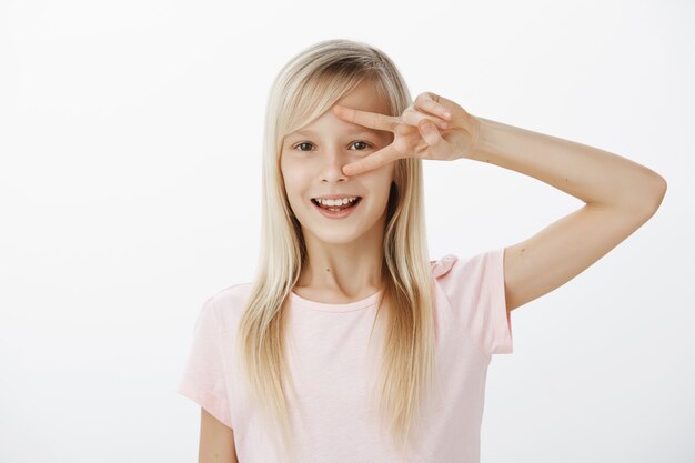 adorabile ragazza bionda con un atteggiamento amichevole che gioca con gli amici e si diverte, mostrando la vittoria o il segno della pace sugli occhi e sorridendo ampiamente dalle emozioni positive sul muro grigio