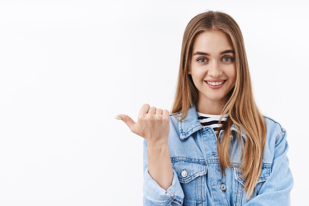 Adorabile ragazza bionda in giacca di jeans che punta a sinistra con il pollice invitante controlla il nuovo merchandising o il prodotto consiglia il miglior negozio di corsi di formazione online stand sfondo bianco sorridente