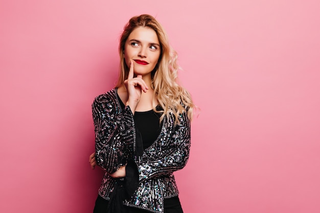 Adorabile ragazza bionda in posa con espressione del viso pensieroso. Piuttosto giovane donna in giacca scintillante in piedi sul muro rosa.