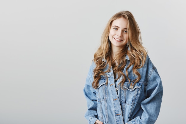 Adorabile ragazza bionda sorridente felice in giacca di jeans, in piedi sopra il muro grigio