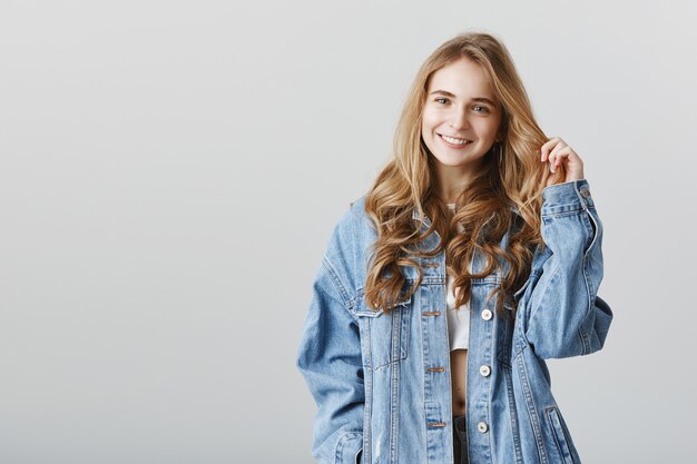 Adorabile ragazza bionda sorridente felice in giacca di jeans, in piedi sopra il muro grigio