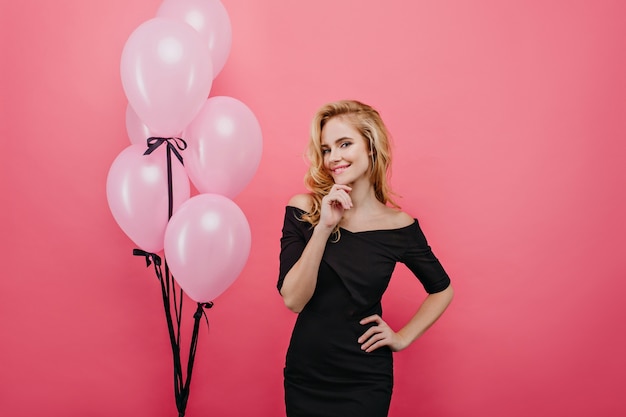 Adorabile ragazza carina con capelli ondulati biondi in posa per il suo compleanno. Ritratto di piacevole giovane donna in nero isolato sulla parete rosa.